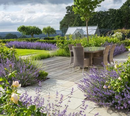 Table and chairs on decking, Nepeta racemosa 'Walker's Low' and Rosa 'Wollerton Old Hall' in borders, view to greenhouse